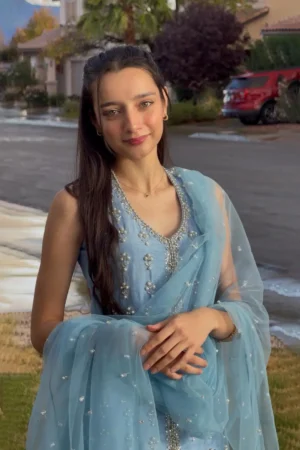 Full-length view of an elegant, light-colored (e.g., ivory or pale gold) raw silk sharara set. The outfit includes a sophisticated long-sleeved top, voluminous sharara trousers, and a sheer tulle dupatta. The ensemble features intricate pearl or zari hand embroidery, reflecting the luxury South Asian artistry of Summer Sekhon.
